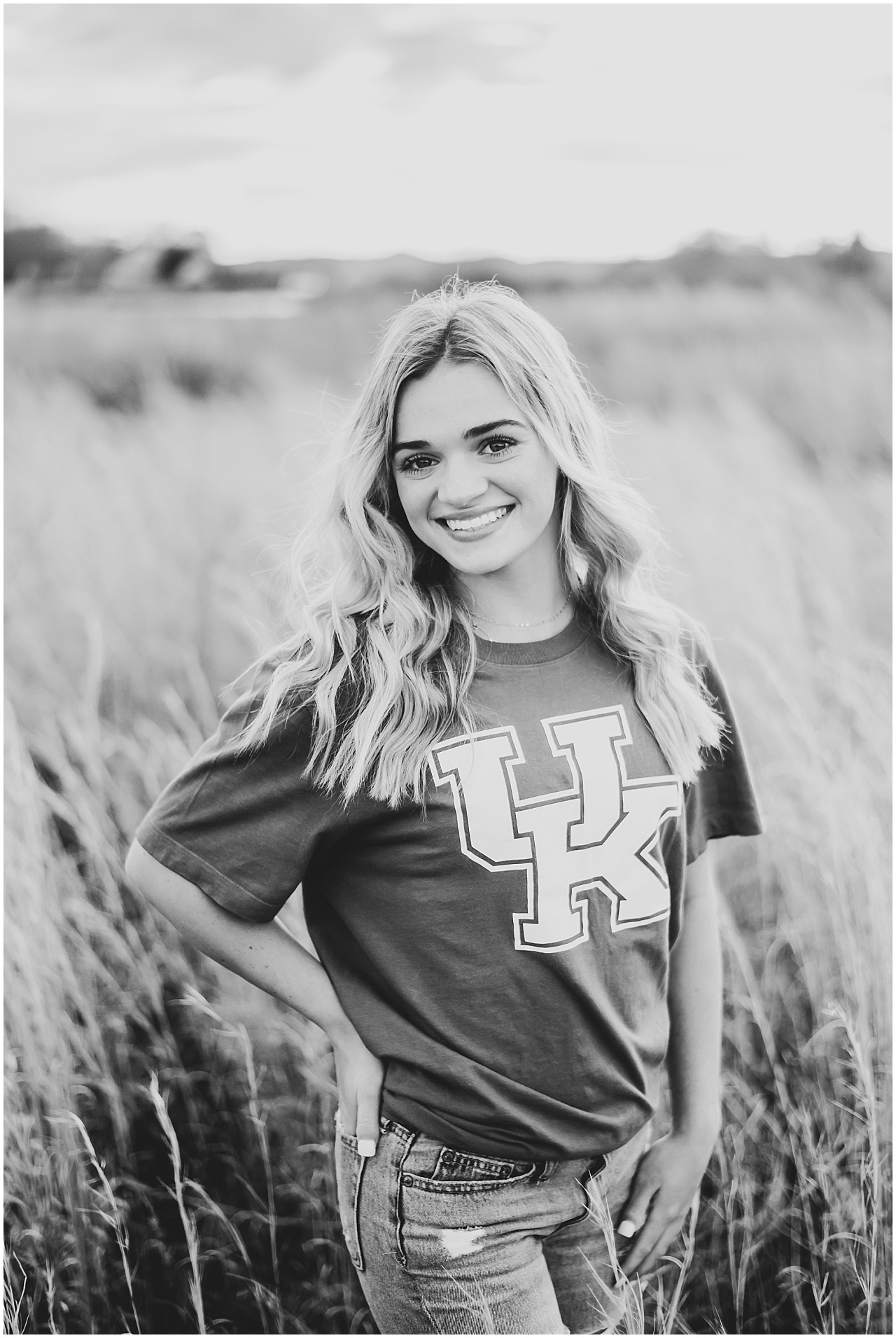 Black and white image of a blonde girl in a field wearing University of Kentucky tshirt and jeans and burkes.